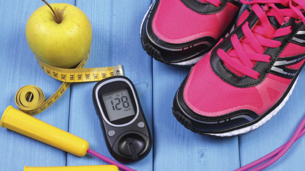 Pink and black sneakers, black blood glucose monitor, pink skipping rope with yellow handles, yellow measuring tape and yellow apple on a blue wooden table